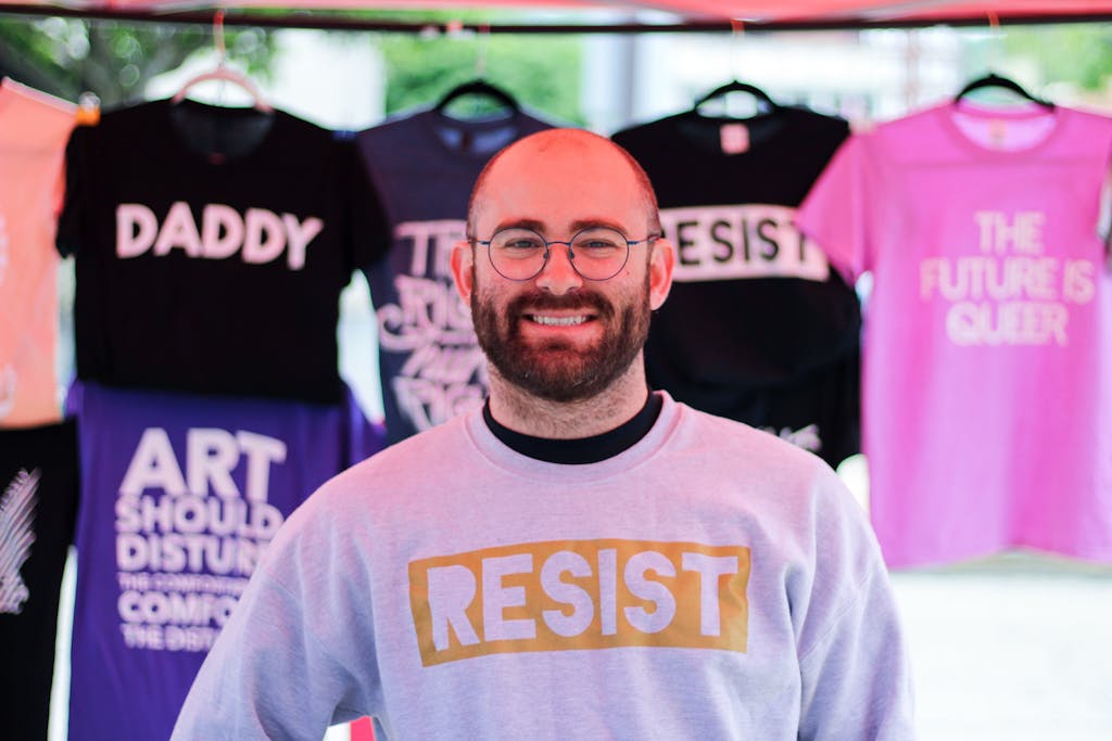 A bearded man wearing a 'Resist' sweatshirt smiles at a market stall showcasing activist-themed shirts.