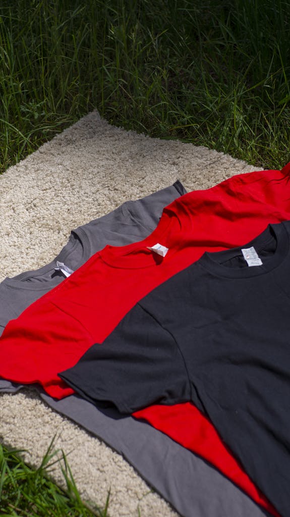 Three vibrant T-shirts in red, black, and grey laid out on a grass field in natural light.